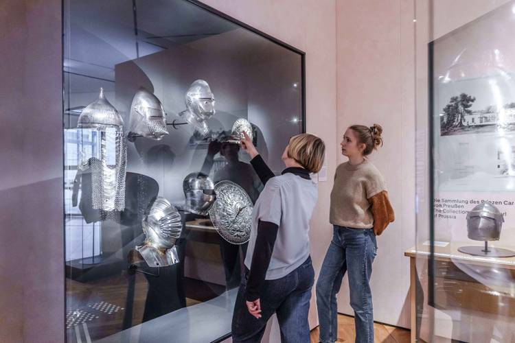 Zwei Besucherinnen stehen vor einer Vitrine. Diese ist an der Wand befestigt. In der Vitrine sind mehrere Helme aus dem Mittelalter zu sehen. Die Besucherinnen beobachten sie.