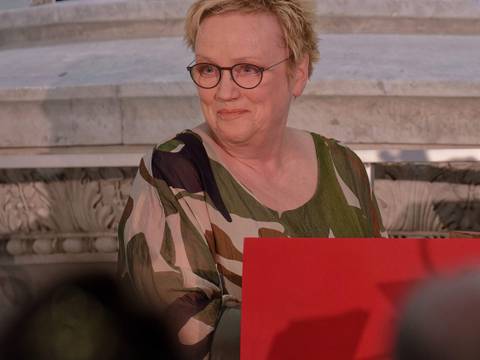  – Eine Person mit kurzen blonden Haaren und Brille sitzt an einem Tisch mit rotem Aufsteller, im Hintergrund Marmordekor.