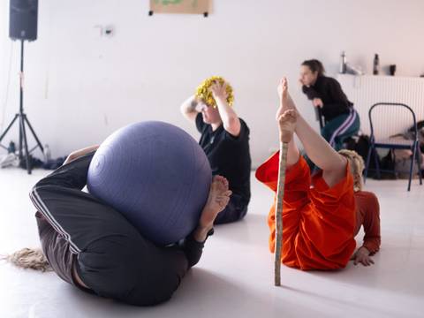  – Drei Personen auf dem Boden in verschiedenen Posen, eine mit einem Gymnastikball, eine mit einem Stock, eine trägt Glitzer auf dem Kopf.