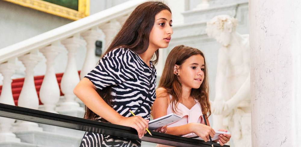 Veranstaltung in der Alten Nationalgalerie – Zwei Kinder auf Treppe in Museum, links Teenager in Zebrakleid, rechts Kind in rosa Top, beide halten Hefte, weiße Statue im Hintergrund.