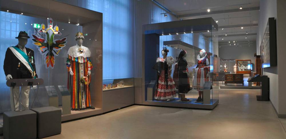 Ansicht der Dauerausstellung „Kulturkontakte. Leben in Europa“ – Ausstellungsraum mit Vitrinen, die bunte traditionelle Trachten an Schaufensterpuppen zeigen. Beleuchtet, hoher Raum, weiße Wände, Glasvitrinen.