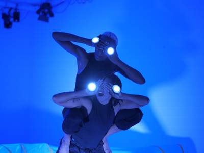  – Der Raum ist in blaues Licht getaucht. Zwei Performerinnen halten sich die Hände vor die Augen. Auf ihren Handrücken befinden sich runde, leuchtende Kreise. Die eine Performerin sitzt aufrecht auf dem Rücken der anderen.
