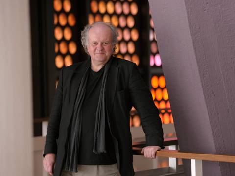 Wolfgang Rihm – Ältere, männlich gelesene Person mit schwarzem Schal und Jacke steht lächelnd an einem Holzgeländer. Hintergrund aus orange leuchtenden Kreisen.
