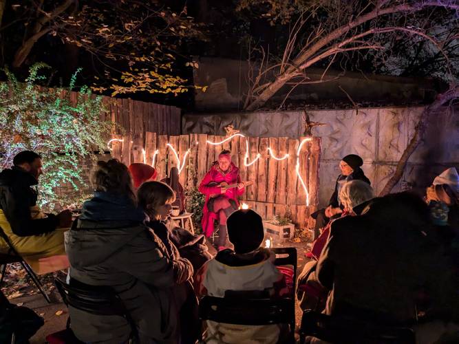 Gruppe von Menschen sitzt im Halbkreis um Lagerfeuer im Garten, dahinter leuchten Lichter am Zaun.	person in roten Kleidern spricht.