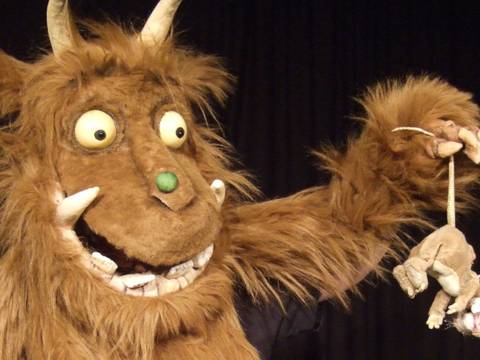 Der Grüffelo – Ein braunes Stoffmonster mit großen Augen und Hörnern hält eine kleine Stoffmaus am Schwanz; schwarzer Hintergrund.