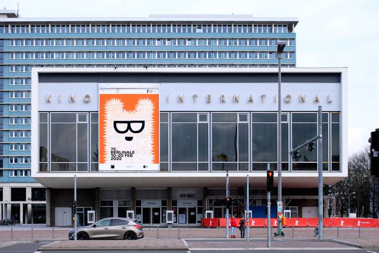 Fotografie „Kino International“, Karl-Marx-Allee, Berlin, 2022 Schriftzug: Klaus Wittkugel (1910–1985), 1961 Berlinale-Plakat: Claudia Schramke (*1985), 2022 – Außenansicht des Kino International in Berlin mit einem großen orange-weißen Berlinale-Plakat, das einen Bären mit Brille zeigt
