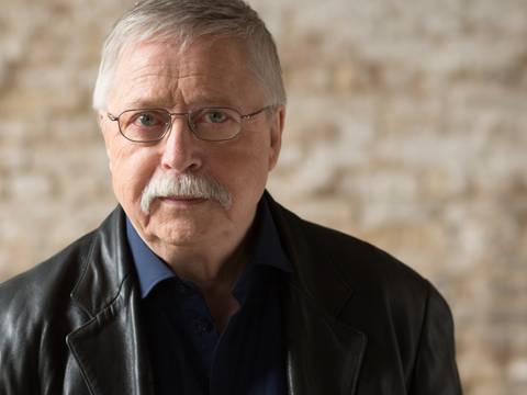 Biermann_Wolf_c_Hans_Scherhaufer_HSM35675.jpg – Eine person mit Brille und grauem Schnurrbart trägt eine schwarze Lederjacke und ein dunkelblaues Hemd vor einer unscharfen Backsteinwand.