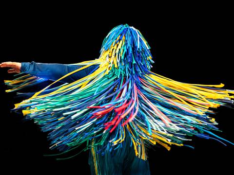 The Sticky Dance | (3-7) & (3-5) – Person mit buntem Fransenanzug in Bewegung, gelbe, blaue und rote Streifen wirbeln um den Körper, schwarzer Hintergrund.