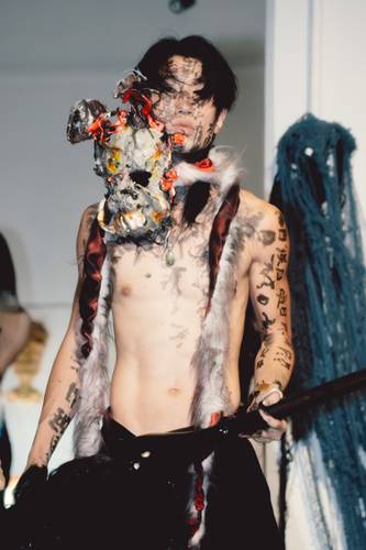 Eine oberkörperfreie Person mit schwarzen Haaren und kalligrafischen Schriftzeichen auf Gesicht und Körper hält ein abstraktes, maskenartiges Objekt vor sich;