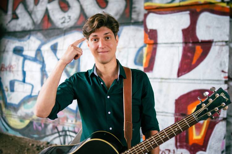 Falk - Unmöglich! – Person zeigt auf den Kopf, lächelt, hält akustische Gitarre; bunte Graffiti an der Wand im Hintergrund.