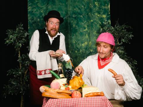  – Zwei Personen an einem Tisch mit Essen, darunter Wurst, Käse und Brot. Eine trägt einen pinken Hut, die andere hält ein Handtuch und schaut fröhlich.
