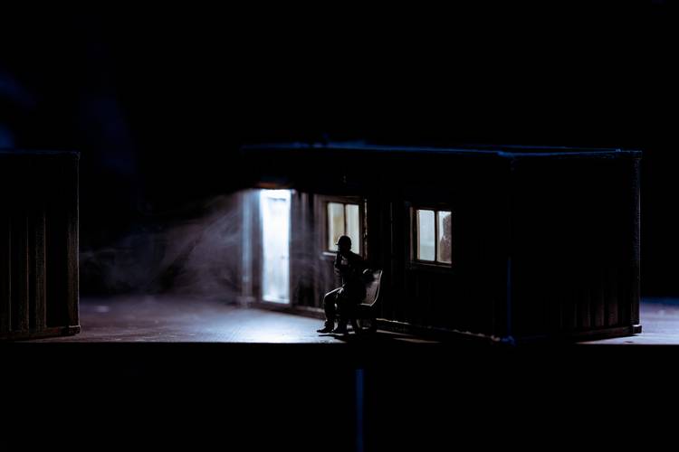 Transport: Tipping Point – Person auf Stuhl vor beleuchtetem Container, lichtdurchflutete Fenster, dunkler Hintergrund, Rauch oder Nebel im Lichtschein.