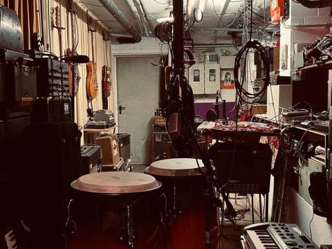 Stroum – Musikraum mit Congas, Keyboards, Kabeln, Verstärkern, Gitarren an der Wand, kleinem Tisch, Regal und Mischpulten. Dezente Beleuchtung.