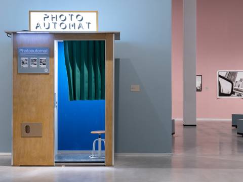 Ausstellungsansicht „Akinbode Akinbiyi. Being, Seeing, Wandering“ – Foto: Ein Foto-Automat mit Verkleidung aus Holz, einem leuchtend blauen Innenraum und einem grünen, geriffelten kurzen Vorhang steht in einem Ausstellungsraum. Im Hintergrund sind Schwarz-Weiß-Fotografien in einem hinteren Raum zu sehen.