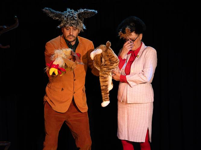 Märchen im Glaspalast von und mit dem Hexenberg Ensemble: Das kleine hässliche Entlein | Bremer Stadtmusikanten – Eine Person trägt Tierohren und hält Handpuppen, während eine zweite Person im Anzug steht und das Gesicht mit der Hand bedeckt.
