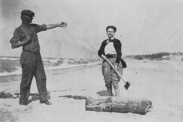 Eine Axt für das gefrorene Meer in uns: Ruth Crawford Seeger, Holzstamm und Freund Carl Sandburg, ca. 1930 – Ein Mann deutet auf eine Frau, die am Strand mit Beil über einem Baumstamm steht.