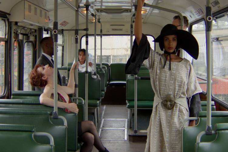 Samuel Chalela Puccini – Person mit schwarzem Hut und hellem Kleid steht in einem Bus und hält sich an der Stange fest. Andere Personen sitzen auf grünen Sitzen.