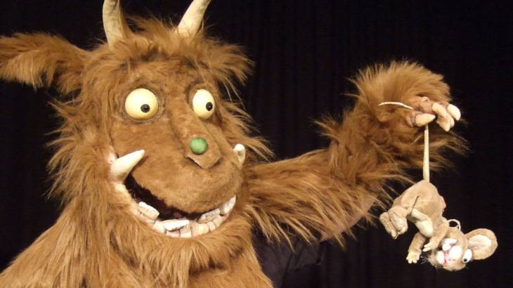 Der Grüffelo – Ein braunes Stoffmonster mit großen Augen und Hörnern hält eine kleine Stoffmaus am Schwanz; schwarzer Hintergrund.