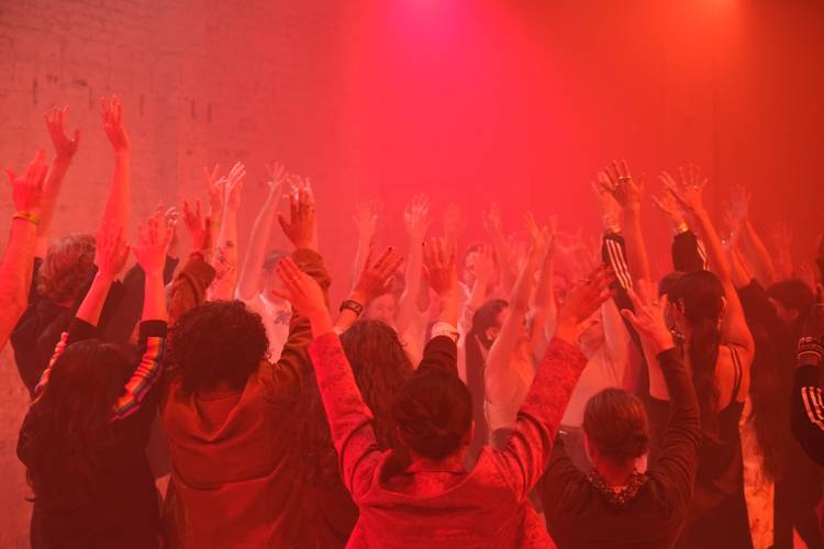 Yoggaton: Transformative Pleasure 2 – Eine Gruppe von Menschen in rotem Licht steht im Kreis und streckt die Arme in die Luft.