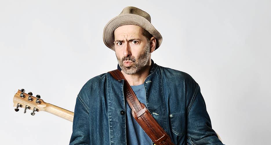 FIL – Mit neuer Show! – Person mit Strohhut und Jeansjacke, Gitarre auf dem Ruecken, schaut mit fragendem Gesichtsausdruck in die Kamera.