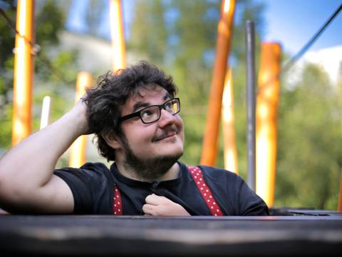 © Dennis Pauls – Mann mit Brille, der sich auf einen Tisch stützt und in die Ferne schaut, mit großen orangefarbenen Stangen im Hintergrund.