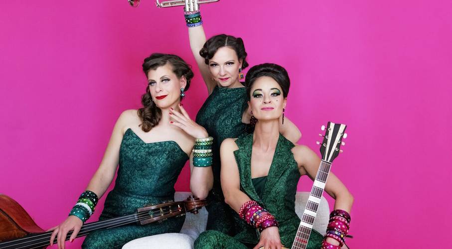 Zucchini Sistaz „Eleganz oder gar nicht!“ – Drei Personen in grünen Outfits vor pinkem Hintergrund. Eine hält eine Trompete, die anderen zwei halten jeweils einen Kontrabass und eine Gitarre.