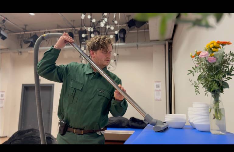Person in grünem Arbeitsanzug mit "Matthäus" Namensschild saugt Staub von blauer Fläche neben Blumenstrauß und weißen Schalen.