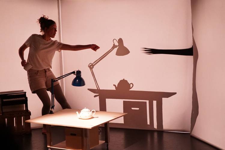 Schattenwerfer – Person zeigt mit ausgestrecktem Arm auf eine Wand, auf der eine Schattenszene mit Schreibtischlampe, Teekanne, Tisch, Arm und Hand projiziert ist.