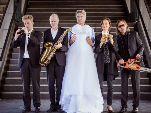  – Fünf Personen in Anzuegen vor Treppe; eine trägt ein Hochzeitskleid, andere halten Fernglas, Saxophon, Bierkrug, Motorsäge.