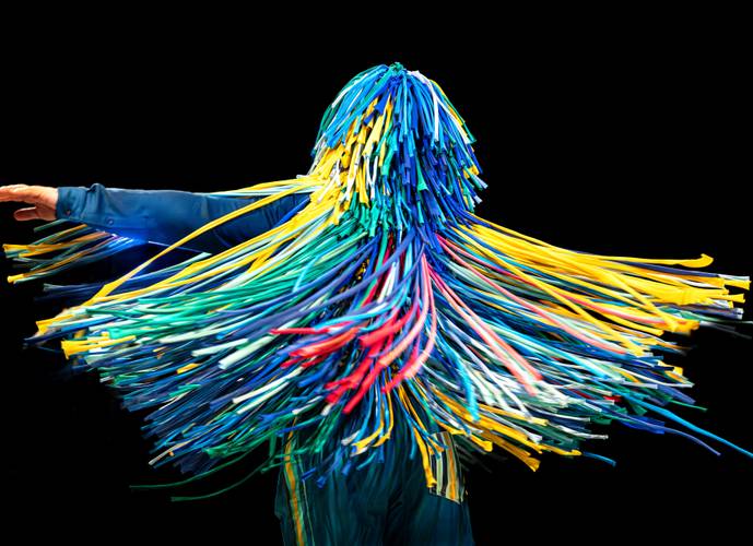 The Sticky Dance – Person mit buntem Fransenanzug in Blau, Gelb, Grün, Rot in Bewegung, schwarzer Hintergrund.