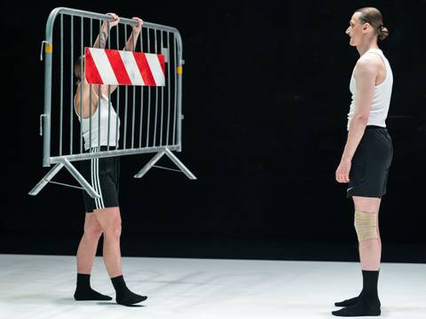 Tanz im August: CLAP & SLAP 1 – Zwei Performer*innen vor schwarzem Hintergrund, eine hält einen Absperrzaun in die Höhe.