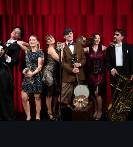 Sechs Personen in Vintage-Kleidung vor rotem Vorhang: mit Gitarre, Saxophon, Mikrofon, Ukulele, Banjo, Tuba. Links stehender Hutträger in Anzug.