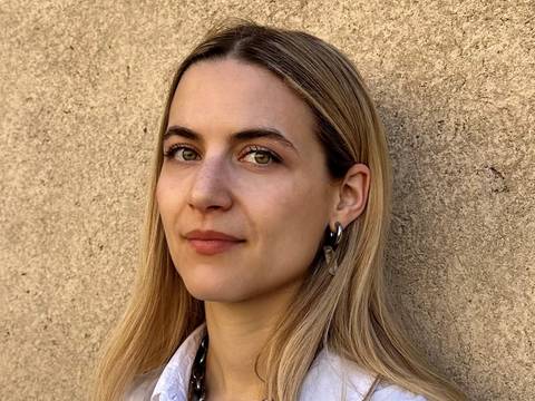 Giulia Giammona – Person mit langem blondem Haar, trägt weiße Bluse und große silberne Halskette, steht vor einer beigen Wand.
