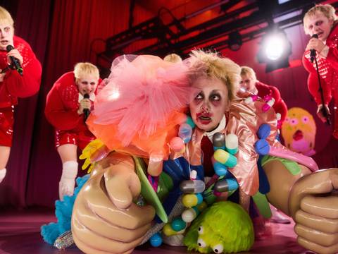 Die Welt im Rücken – Bühnenperformance mit mehreren Personen in glitzernden roten Kostümen; im Vordergrund eine farbenfroh verkleidete Figur mit großen, überdimensionierten Requisitenhänden und buntem Dekor, umgeben von grellem Bühnenlicht.