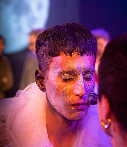 Person mit Glitzer im Gesicht und geschlossenen Augen trägt weißes, durchscheinendes Gewand. Eine andere Person trägt Goldohrring und pinselt das Gesicht.