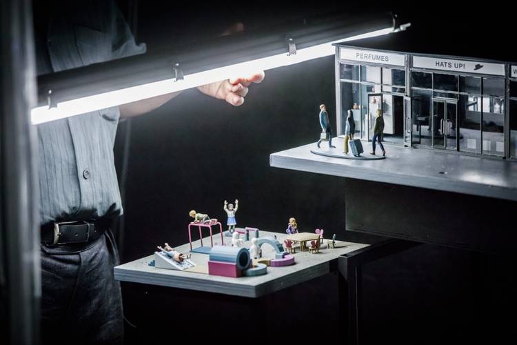 Transport: Connecting Flights – Miniaturszene mit Figuren an einer Eingangstür mit Text "PERFUMES HATS UP!". Hand justiert Beleuchtung. Darunter Spielplatz mit Kindern.