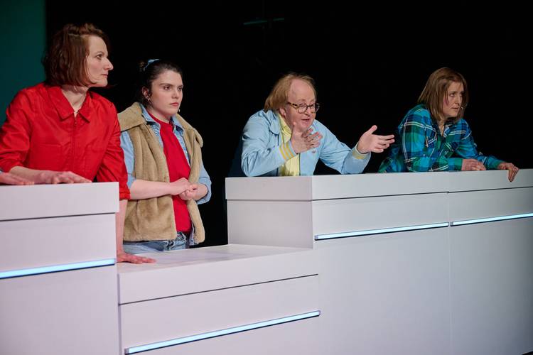 Vier Personen an weißen Podesten, links rote Jacke, daneben rote Weste, Person in der Mitte gestikuliert, rechts karierte Jacke. Schwarz-grauer Hintergrund.