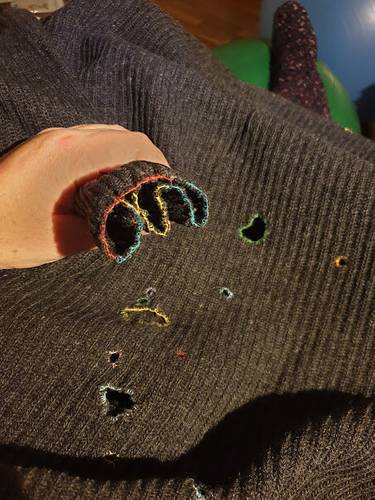 Eine Hand im grauen Strickpullover mit bunten Saumkanten. Mehrere sichtbare Loecher im Pullover. Im Hintergrund ein gruener und ein blauer Gegenstand.