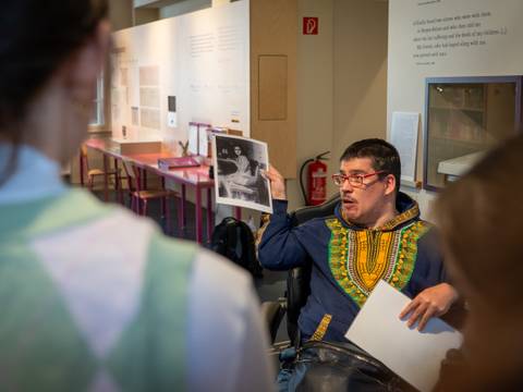 Peer Guides begleiten durch die Ausstellung – Mann in Ausstellung zeigt einer Gruppe ein historisches schwarz-weiß-Foto von Anne Frank.