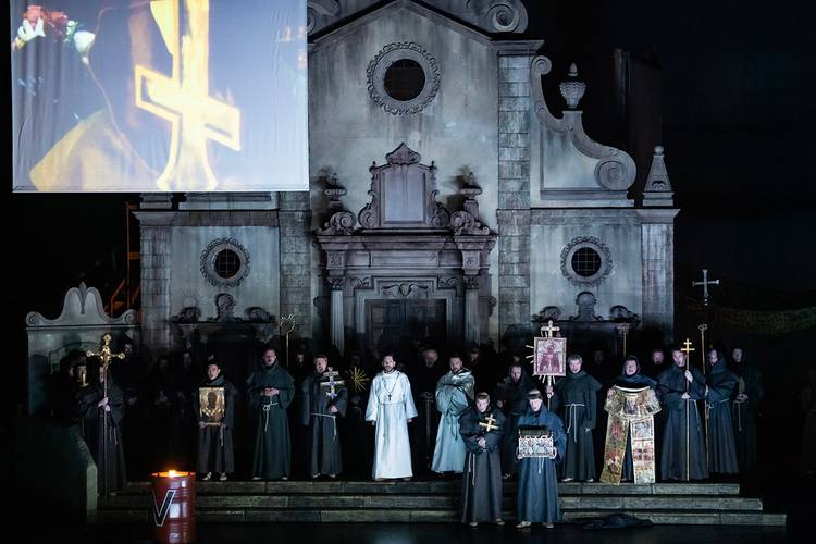 La forza del destino – Mehrere dunkel gekleidete Personen und eine in Weiß vor einer Kirche, halten Kreuze und Ikonen. Großes Bild zeigt gelbes Kreuz auf einer Leinwand.