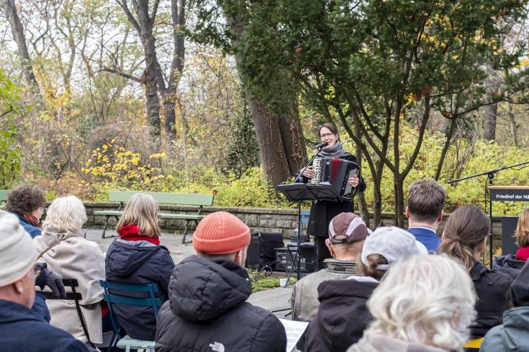 Person spielt Akkordeon vor Publikum im Freien, umgeben von Bäumen. Schild mit Text: Friedhof der Märtyrer von 1848. Herbstliche Atmosphäre.