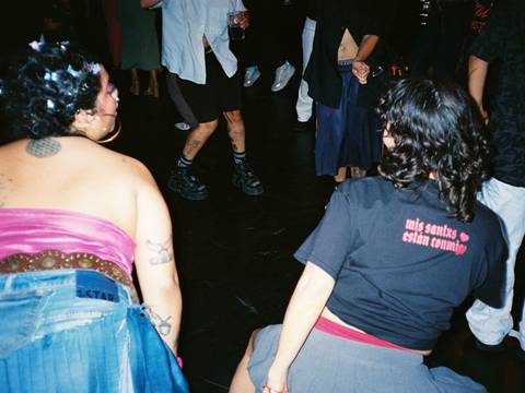 Fotos: Inmensidades – Personen tanzen in einem Club. Eine Person trägt ein T-Shirt mit der Aufschrift "mis santxs estan convmxg".