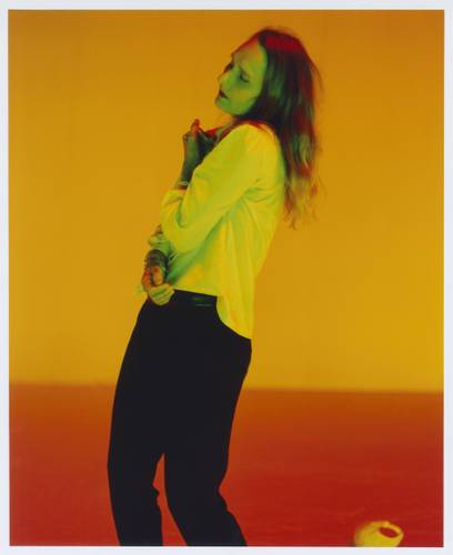 Foto: Estelle Hanania – Langhaarige Person in weißem Hemd und schwarzer Hose vor orange-gelbem Hintergrund, mit geschlossenen Augen und seitlich geneigtem Kopf.