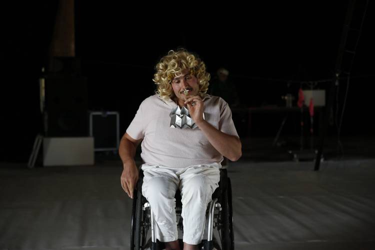 Performer*in mit blonder Lockenperücke sitzt im Rollstuhl auf dunkler Bühne und hält eine kleine Blume an die Nase.