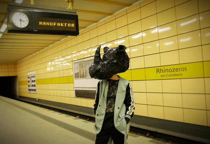 Rhinozeros – ein Comeback – Person mit schwarzem Nashornkopf in U-Bahn-Station. Auf Schriftzug steht "Rhinozéros ein Comeback". Wandkacheln gelb. Uhr zeigt 10:10, Anzeigetafel