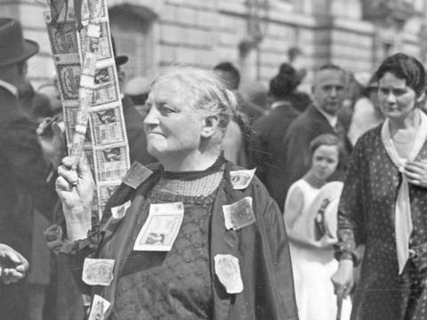 Fotografie „Eine Rentnerin protestiert mit aufgenähten Banknoten gegen die Geldentwertung“ – Schwarzweißfoto mit älterer Person, die ein Schild mit Bildern und Text haelt. Kleidung mit Geldscheinen. Menschenmenge im Hintergrund.