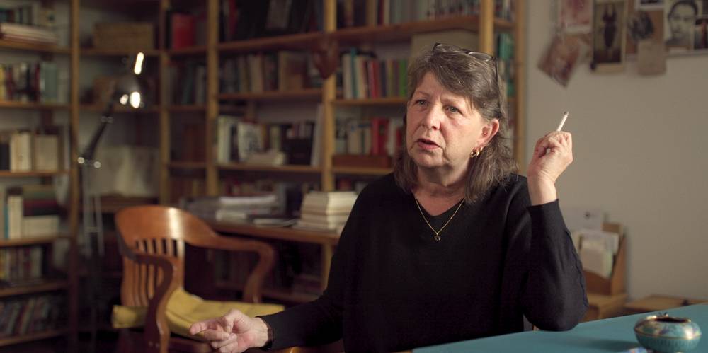 Helene Weigel - Revolutionärin im Rampenlicht – Ältere Person mit dunklem Pullover sitzt an einem Tisch, spricht, hält eine Zigarette. Bücherregale und ein Stuhl im Hintergrund.