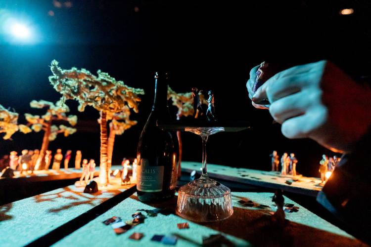 Transport: Fasten your Seatbelts! – Miniaturfiguren auf einem Glas; Hintergrund mit Bäumen. Prosecco-Flasche mit Etikett "Spumante" links. Hand platziert Figur auf dem Glasrand.