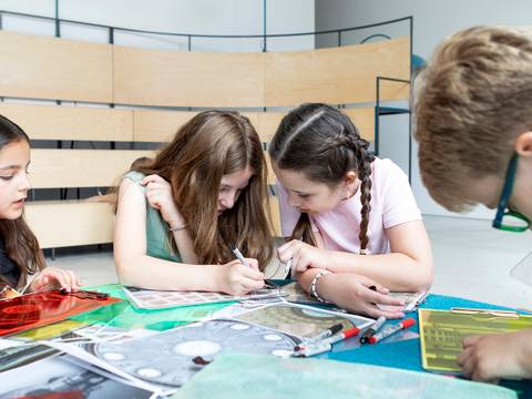  – Fünf Kinder sitzen an einem Tisch und sind in eine kreative Aktivität vertieft, mit Papieren, Stiften und durchsichtigen Farbfolien.