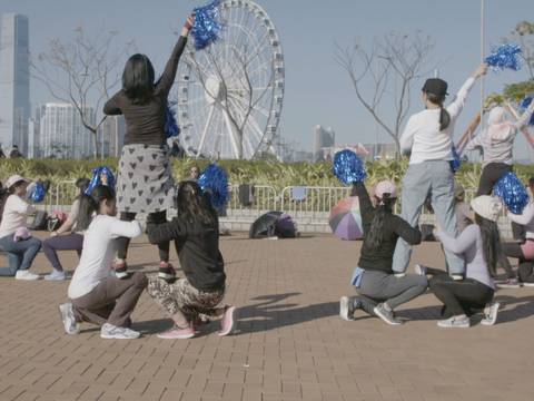 Stephanie Comilang, Come to Me, Paradise, 2017 – Video-Still: Eine Gruppe von Menschen im Freien mit blauen Pompons, einige kniend und andere stehend, vor einem Riesenrad und Hochhäusern in einer Stadtlandschaft.